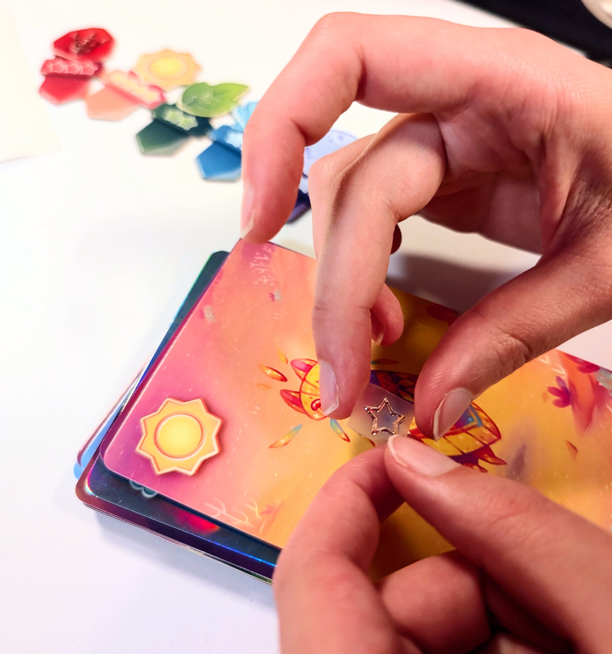 Deux mains déposent un petit jeton en forme d'étoile sur une carte colorée ornée d'un symbole représentant un soleil. D'autres cartes et jetons colorés sont éparpillés sur une surface blanche à l'arrière-plan.