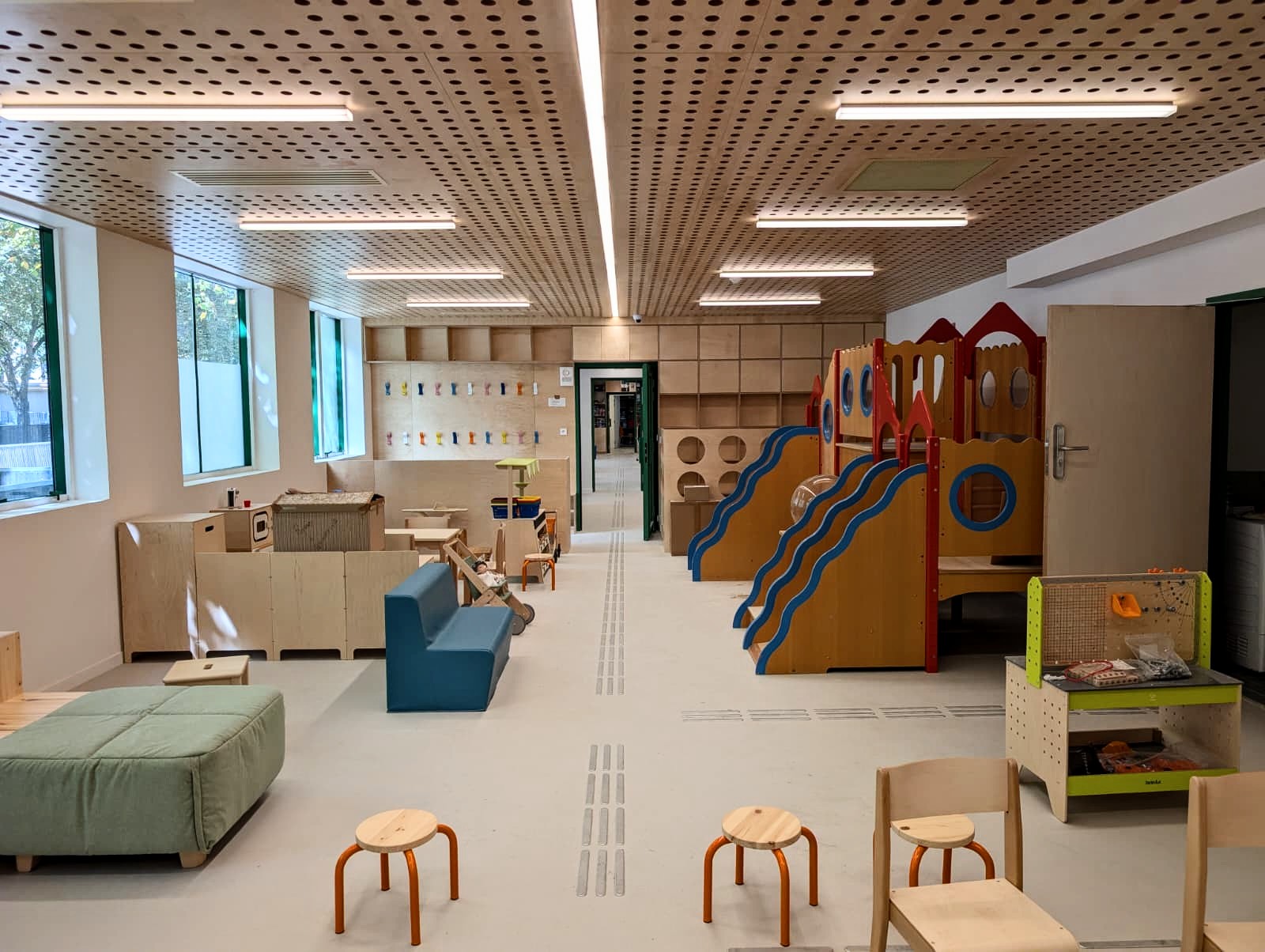 Une salle de classe de maternelle lumineuse, équipée de mobilier en bois, d’aires de jeux d’intérieur colorées avec des toboggans, de petites chaises, d’étagères, de livres et de grandes fenêtres laissant entrer la lumière naturelle.